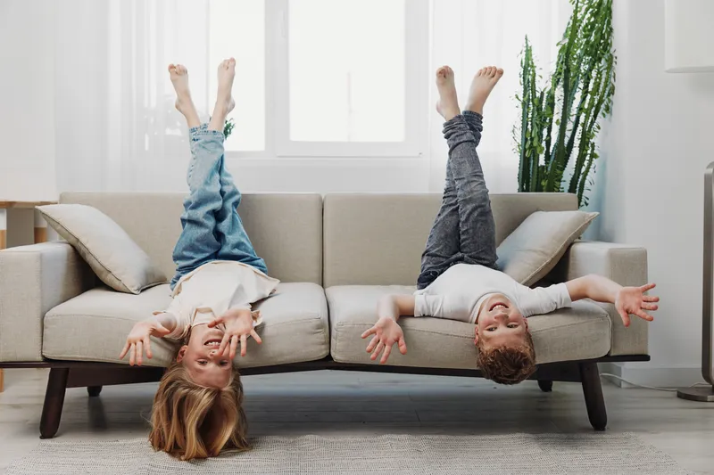 Two happy children wearing casual clothes lie upside down on a beige sofa in a bright living room with a large window. Concept of playfulness and family fun.