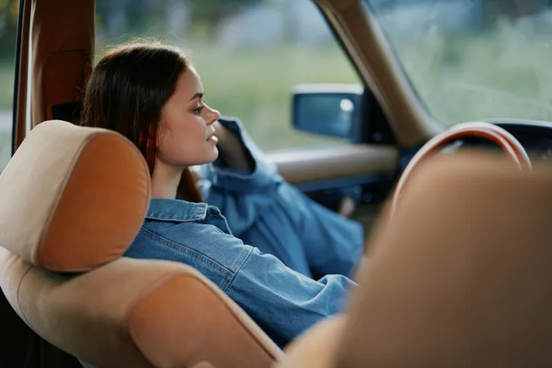 Young woman in a denim jacket sitting relaxed in a vintage car, reflecting calm emotions, featuring warm tones and a soft background.