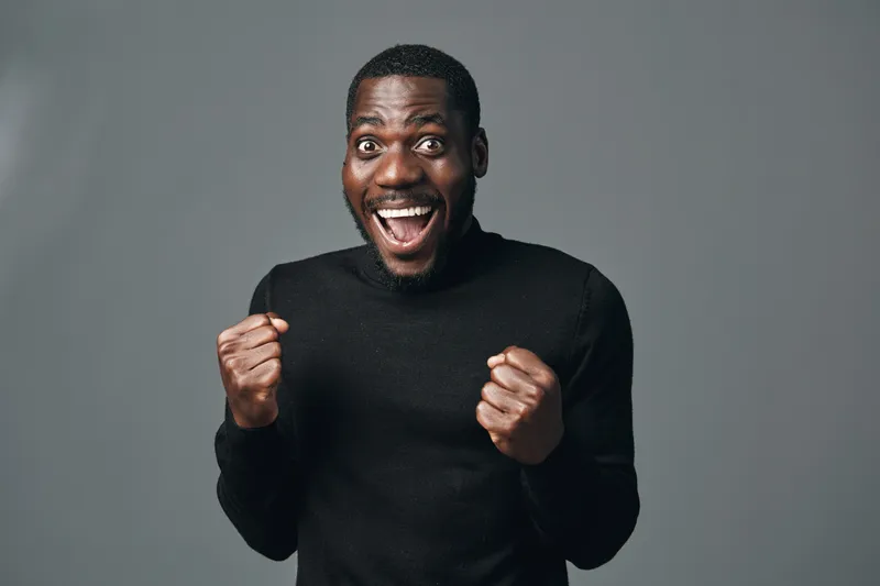 African man with dark skin wearing black sweater showing excitement and joy with clenched fists and wide smile in creative conceptual visual style studio portrait.