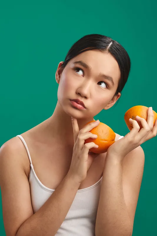 asian woman holds bright oranges against a vivid green backdrop, wearing a light top. Her thoughtful gaze and soft lighting create a fresh, natural lifestyle vibe.