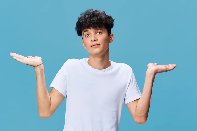 Young man shrugging with open palms wearing a white t-shirt on blue background, expressing uncertainty or questioning with casual hairstyle.