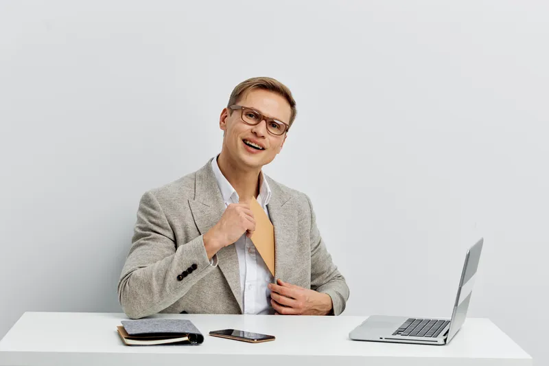 Smiling man in business casual attire sitting at desk with open laptop, holding a yellow folder, isolated on plain white background. Modern workplace concept, professional, happy, male