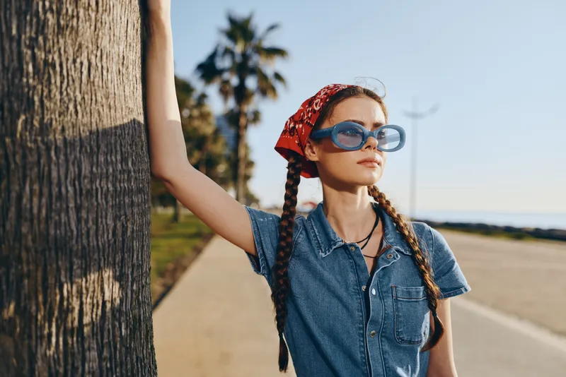 Lifestyle woman in modern boho-western denim street style wearing sunglasses and a red bandana, captured with warm film color tones outdoors in sunny weather.