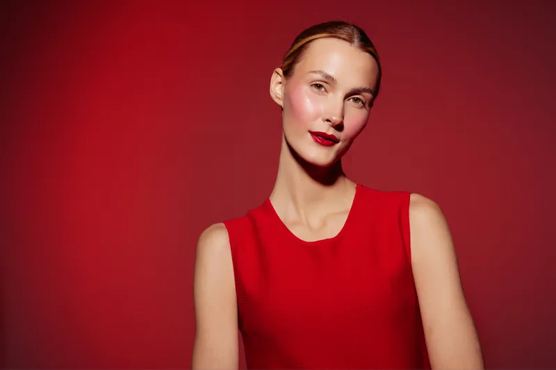 Portrait of a confident woman in a red sleeveless dress against a red background. She has sleek hair and red lipstick, creating a bold and elegant fashion concept.
