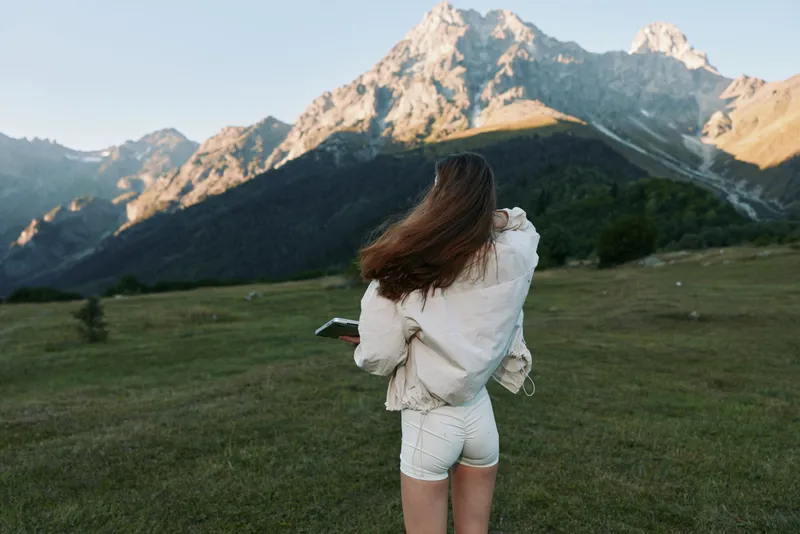 A young woman stands in a wide meadow facing towering mountains, wearing a light jacket and shorts, holding a tablet and admiring the scenic landscape.