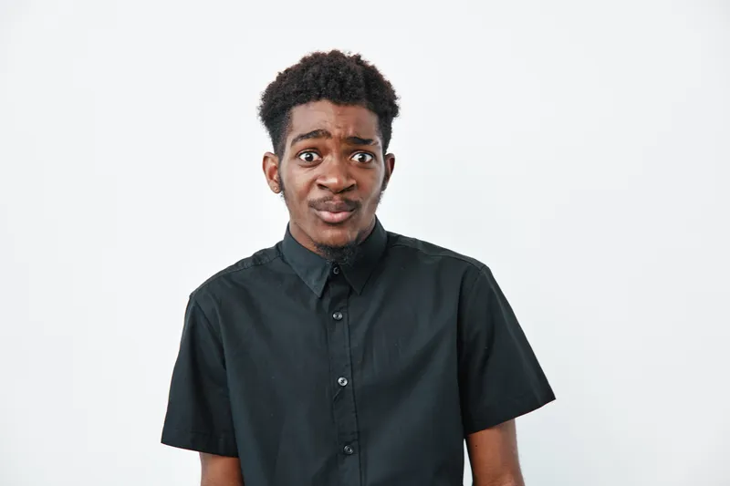 Young black man with dark skin and curly hair wearing black shirt looking confused or skeptical on white background. African ethnicity male expressing doubt or uncertainty in studio.