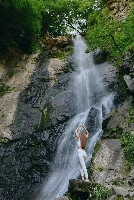 woman standing by a tall waterfall surrounded by rocks and lush green trees, wearing white leggings, stretching with arms raised under natural daylight in forest setting