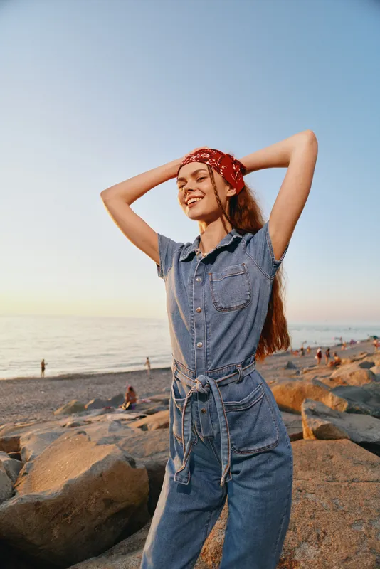 Lifestyle woman in modern boho-western denim street style enjoys seaside sunset with relaxed smile and casual pose, film color tones enhance warm natural light.