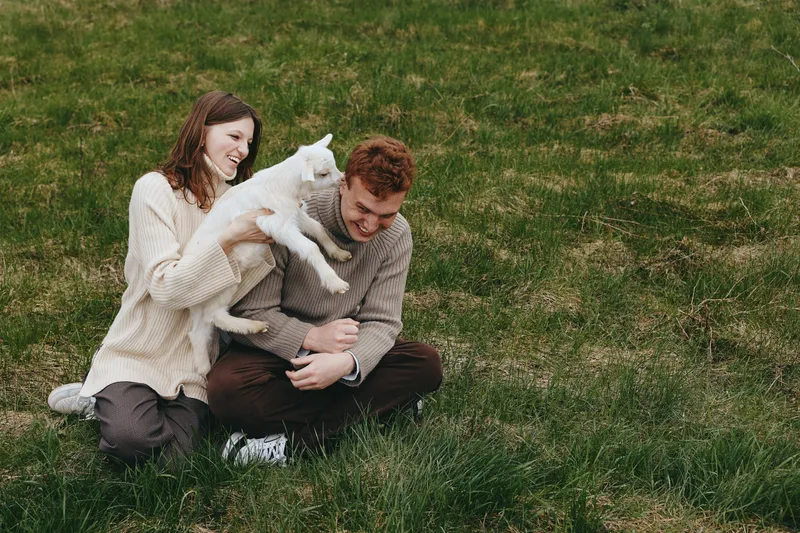 Playful couple on a grassy field enjoying time with a friendly goat kid, capturing warm interaction and bonding in a casual outdoor moment. Natural daylight enhances a relaxed, joyful mood.