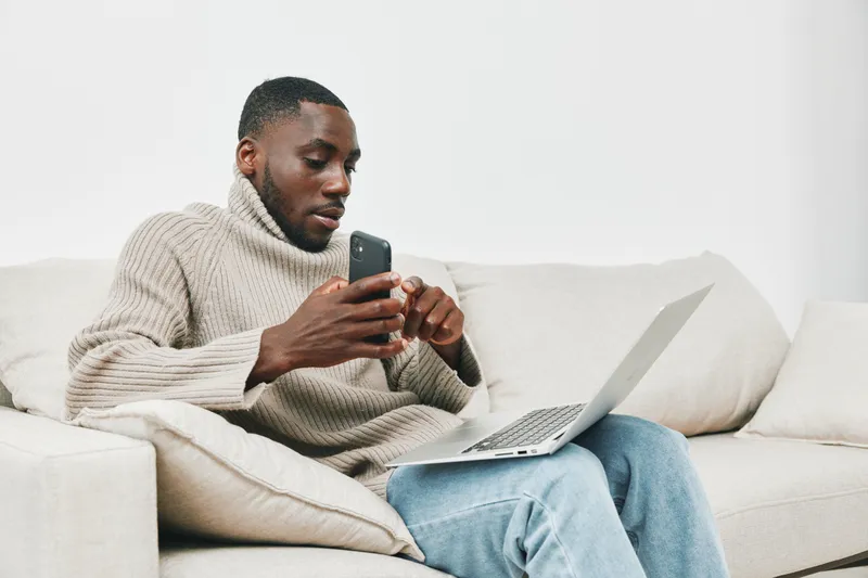 Young African man in cozy knit sweater engaging with a smartphone while using a laptop, showcasing a modern and productive workspace atmosphere