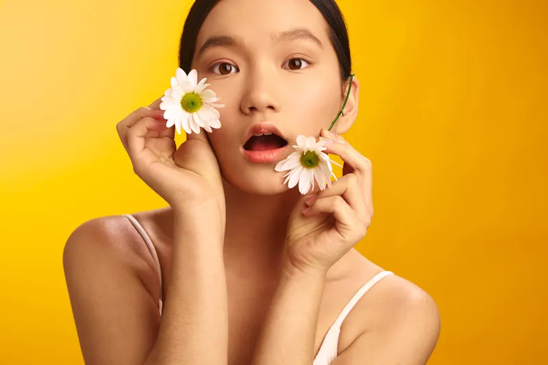 Asian woman holding white daisies near her cheeks against a vivid yellow backdrop, with a gentle, surprised expression and playful mood that conveys fresh beauty and optimism.