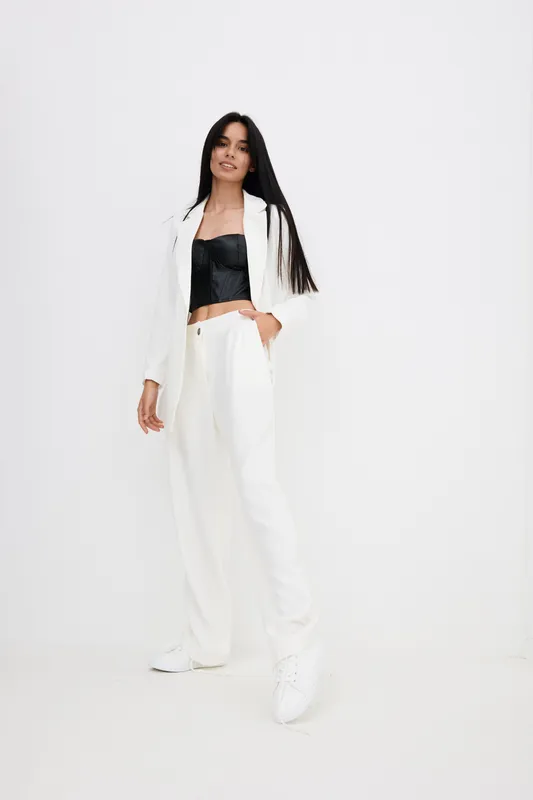 Tall woman in white suit and black top posing confidently against plain white background, showcasing modern fashion style and relaxed posture.