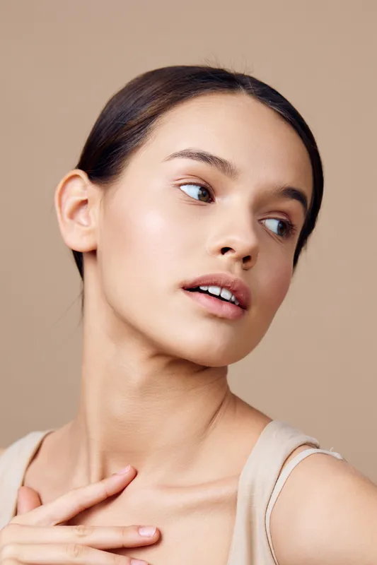 Natural beauty portrait of a young woman with a soft expression, wearing a neutral toned outfit, against a light beige background, highlighting healthy skin and serenity