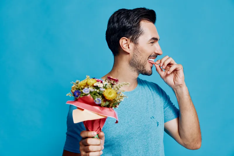 Smiling man holding a bouquet of flowers while playfully biting his finger, dressed in a casual blue t-shirt against a vibrant blue background. Joyful lifestyle concept.