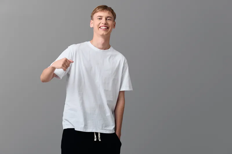 Thumbs up from a casual young man wearing a white t shirt, smiling against a gray studio backdrop, conveying friendly approval and relaxed everyday style, positive energy.