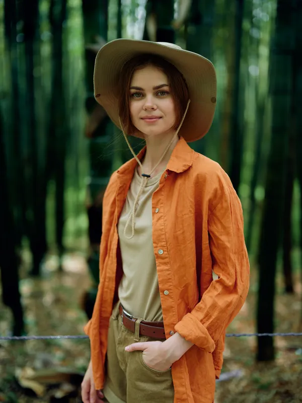 Woman portrait outdoors in a bamboo forest wearing a wide hat and orange jacket, casual traveler pose with relaxed smile and natural light, green background for lifestyle photography.