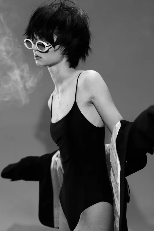 Black and white portrait of a stylish woman wearing sunglasses and a black swimsuit with moody atmosphere and smoky background
