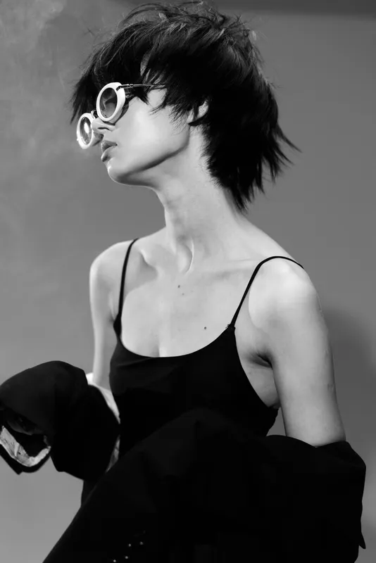 Black and white portrait of a young woman with short hair wearing round glasses and a black outfit, showing a stylish and contemplative mood.