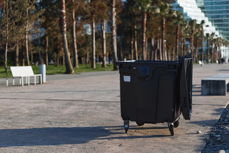 Black outdoor trash bin on wheels standing on a paved path in a park with trees and benches in the background. Urban waste management concept in daylight.