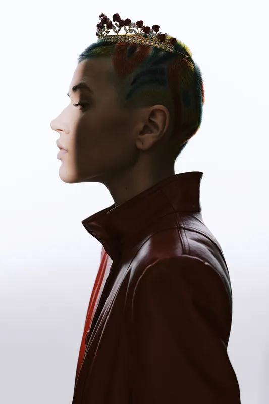 Profile of a woman with short rainbow-dyed hair wearing a decorative crown and a leather jacket, featuring a modern edgy look in a minimalist high-key studio setting.