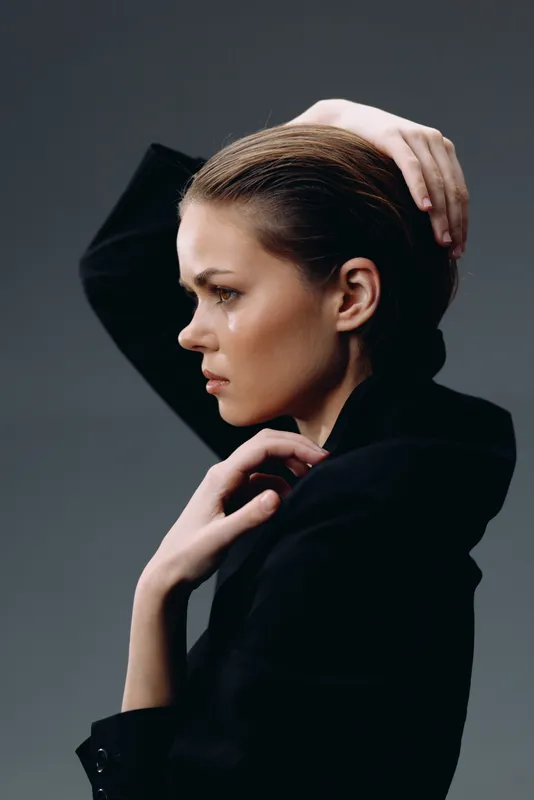 Profile of a thoughtful woman with slicked-back hair in a black blazer against a dark background.