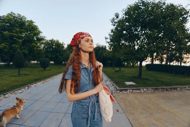 Lifestyle woman in modern boho-western denim street style stands outdoors with a red bandana, long hair, and a casual tote bag in natural light film color tones.