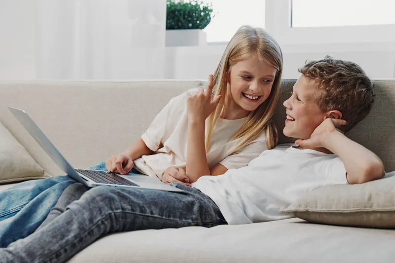 Happy children, boy and girl, relaxed on couch using laptop, smiling and interacting in bright living room. Concept of family, technology, leisure, and childhood joy.