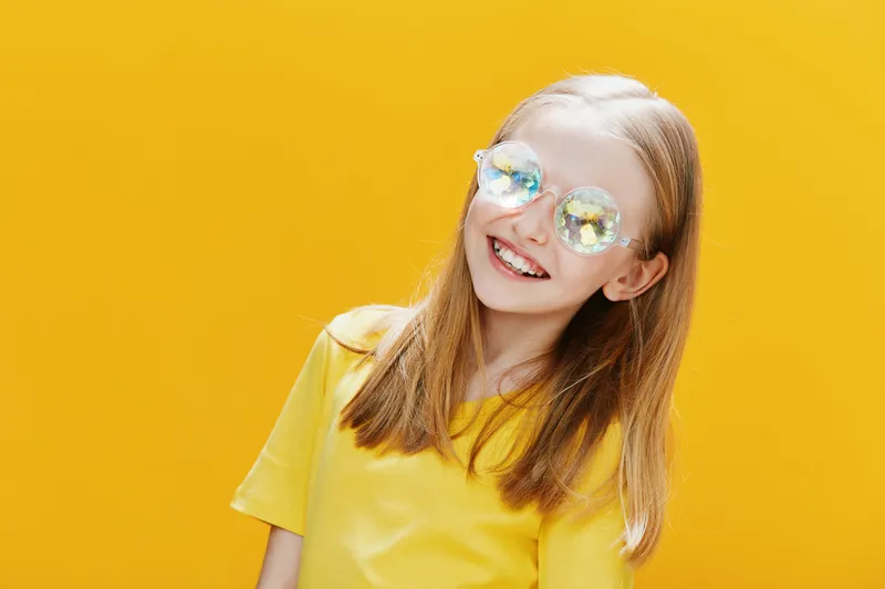 Cheerful girl with long hair wearing stylish sunglasses and a yellow shirt, smiling against a vibrant yellow background, reflecting joy and summer vibes