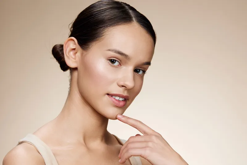 Young woman with smooth skin and a gentle smile, showcasing healthy beauty and confidence against a soft beige background, emphasizing natural radiance and femininity