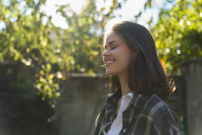 Brunette woman with shoulder length hair smiles softly in sunlit outdoors, wearing a plaid shirt over a white top, conveying a casual, relaxed mood and everyday contentment