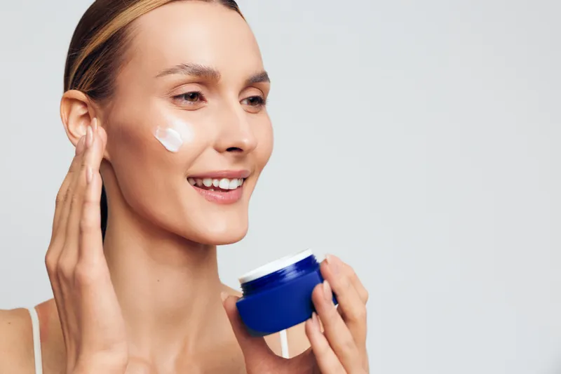 Young woman applying face cream with smile, healthy skin care routine, natural makeup, soft lighting, white background, close up portrait, beauty concept