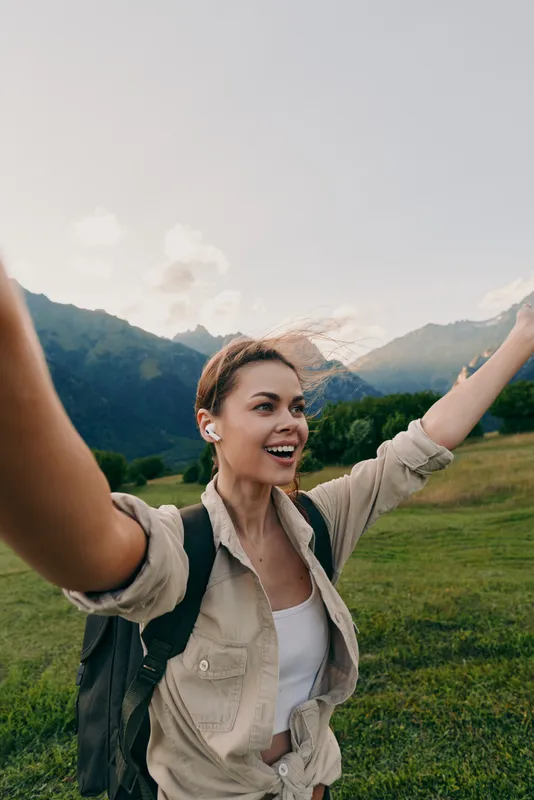 Happy woman hiking outdoors with backpack and earbuds, smiling with arms raised, mountain landscape background, casual outfit, natural light, active lifestyle