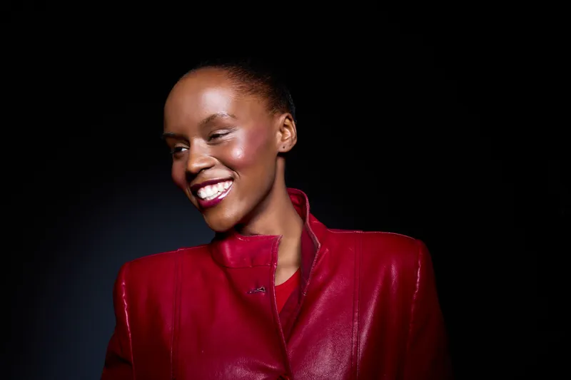 Smiling black woman with dark skin wearing a red leather jacket looks away from the camera on a dark background. Portrait of happy African American female model with natural makeup.
