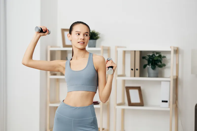 Female fitness enthusiast performing shoulder press with dumbbells in a bright home gym, wearing athletic activewear, showing strength, balance and focus during a workout session