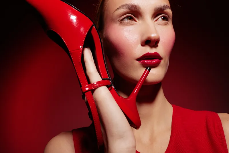 Close-up of a stylish woman with red lipstick holding a red high heel shoe near her face. The red background and dress create a bold fashion concept with strong color contrast.