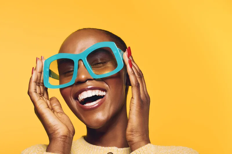 Happy dark skinned African American woman with shaved head wearing oversized blue glasses smiling with closed eyes on yellow background. Concept of joy, fashion, and beauty.