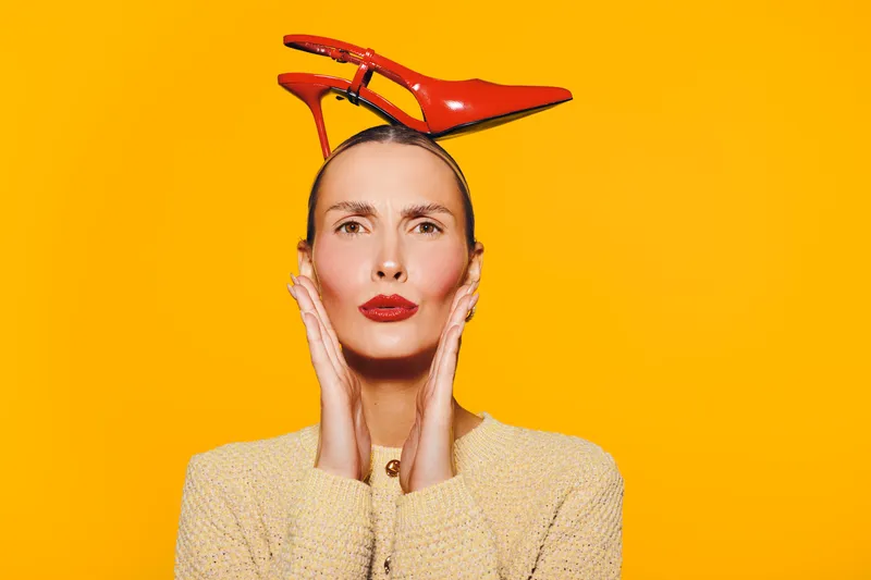 Stylish woman with red high heel shoes balanced on her head poses against bright yellow background. Concept of fashion, creativity, and bold style expression in studio lighting.