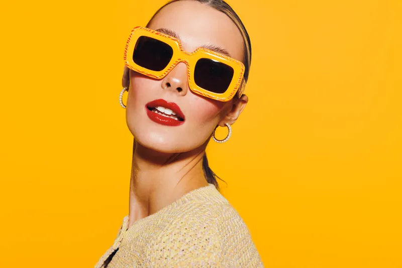 Stylish woman wearing large orange sunglasses and red lipstick poses confidently against bright yellow background. Fashion concept with bold colors and modern accessories.