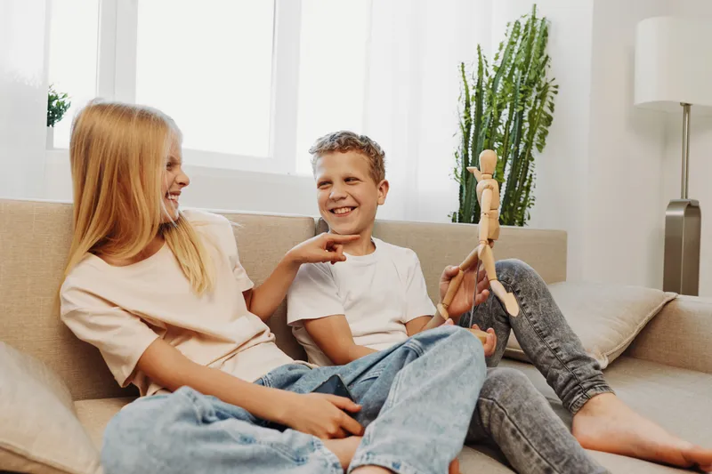 Two happy children sitting on a beige sofa in a bright living room, smiling and playing with a wooden mannequin. Casual clothes and natural light create a warm, playful atmosphere.