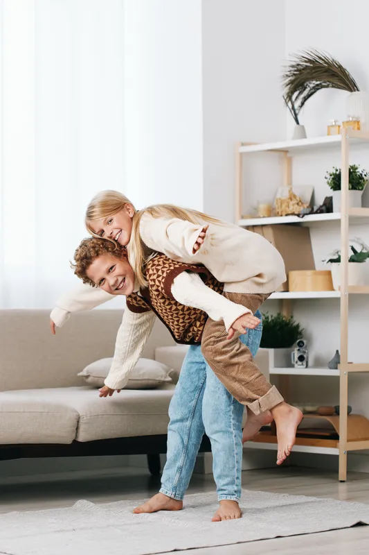 A playful moment as a father carries his child on his back indoors, showcasing balance, laughter, and a cozy living room scene with warm natural light.