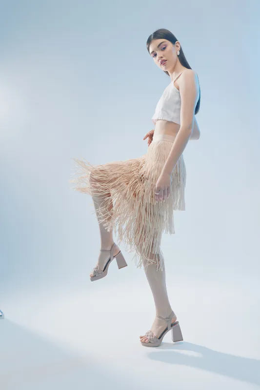 Slim woman in stylish beige fringe skirt and white crop top poses with one leg raised on a light blue background. Concept for woman beauty fashion and elegance.