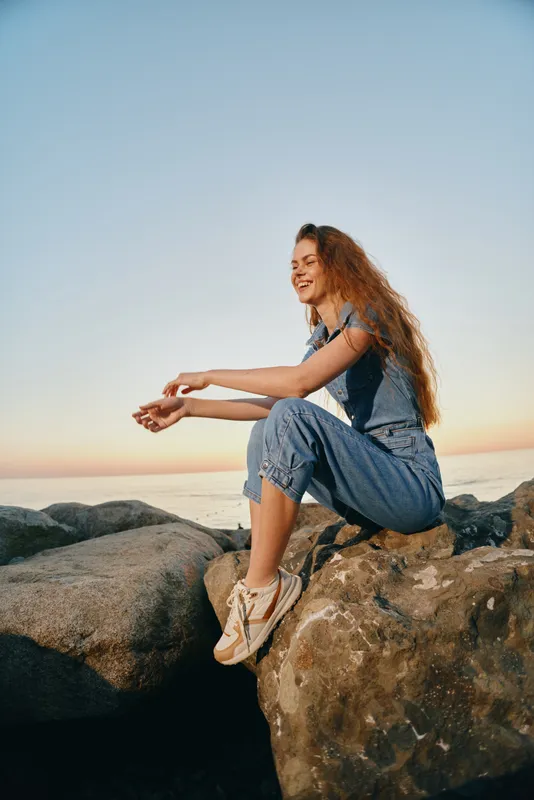 Lifestyle woman in modern boho-western denim street style sitting on rocks, smiling with natural red hair and casual sneakers, film color ambiance at sunset.