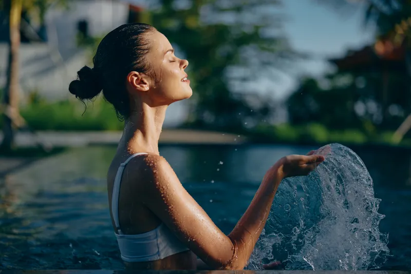 Young woman in white swimsuit enjoys refreshing water splash in outdoor pool on sunny day with closed eyes and relaxed face, lifestyle concept of leisure and wellness.