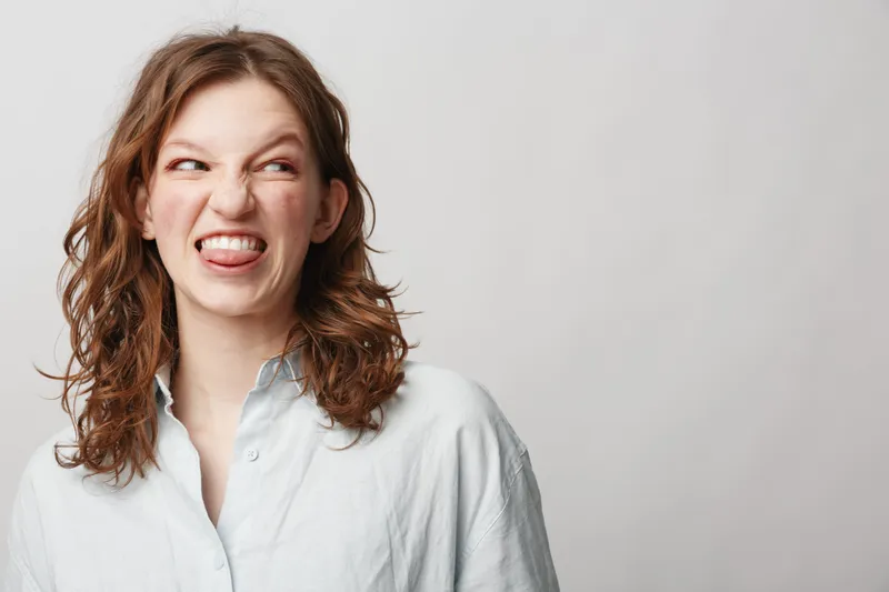 Young woman with curly hair makes funny face showing tongue and scrunched eyes. Casual white shirt and neutral background. Concept of humor, expression, and personality.