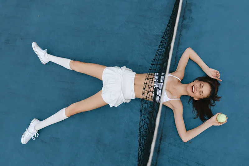 Happy female athlete lying on a tennis court, wearing white sportswear, smiling while holding a tennis ball, showcasing a carefree and sporty vibe