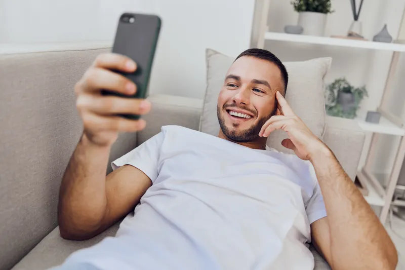 Happy man lying on a couch, smiling at his smartphone, enjoying a relaxing moment at home. Casual setting with natural light and modern decor.