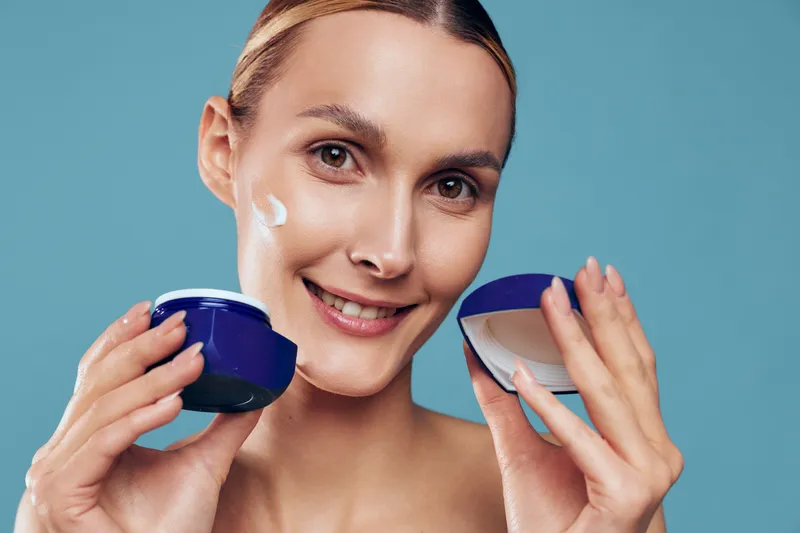 Young woman applies moisturizing cream on face holds open jar with white lotion smiles looks at camera isolated on blue background skincare beauty concept.