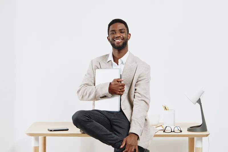 Smiling man in casual business attire sitting at desk holding laptop, with office supplies and computer on background, studio shot, white background, cheerful professional lifestyle concept.