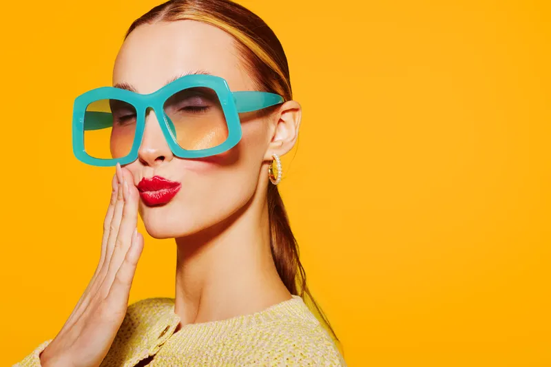 Stylish woman with large turquoise glasses and red lipstick blows a kiss on bright yellow background. Concept of fashion, beauty, and playful attitude in studio portrait.