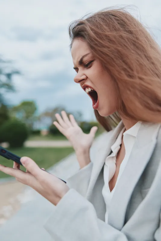 Businesswoman expressing frustration while using smartphone in outdoor setting. Emotionally charged moment with nature background. Stylish, modern attire.